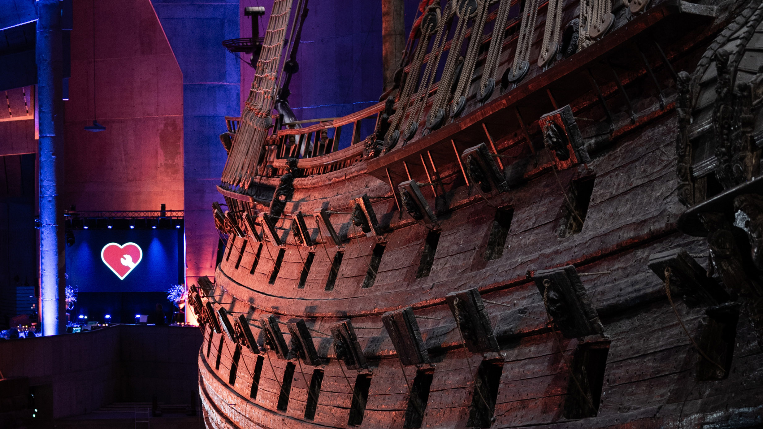 Foto av skeppet Vasa sett snett bakifrån, med en symbol med rött hjärta med vit skiftnyckel på skärm framför skeppet.a