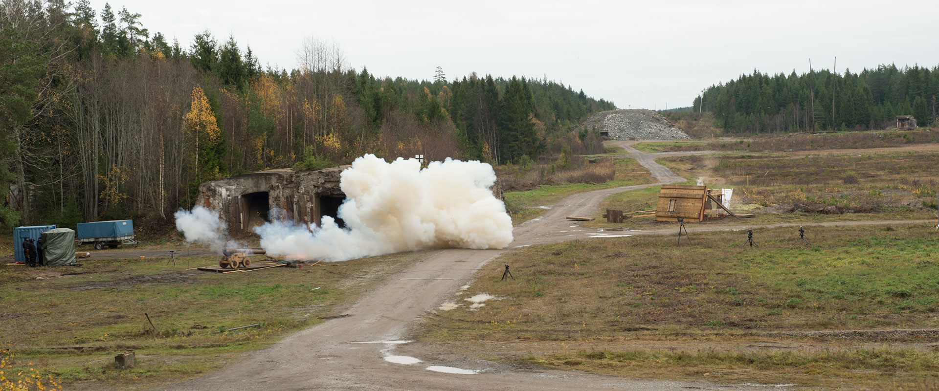 En kopia av en Vasakanon har avfyrats mot en kopia av Vasas skrovsida. Ett moln av krutrök syns framför kanonen. I bakgrunden skymtar skog och i mitten av bilden en grusväg. 